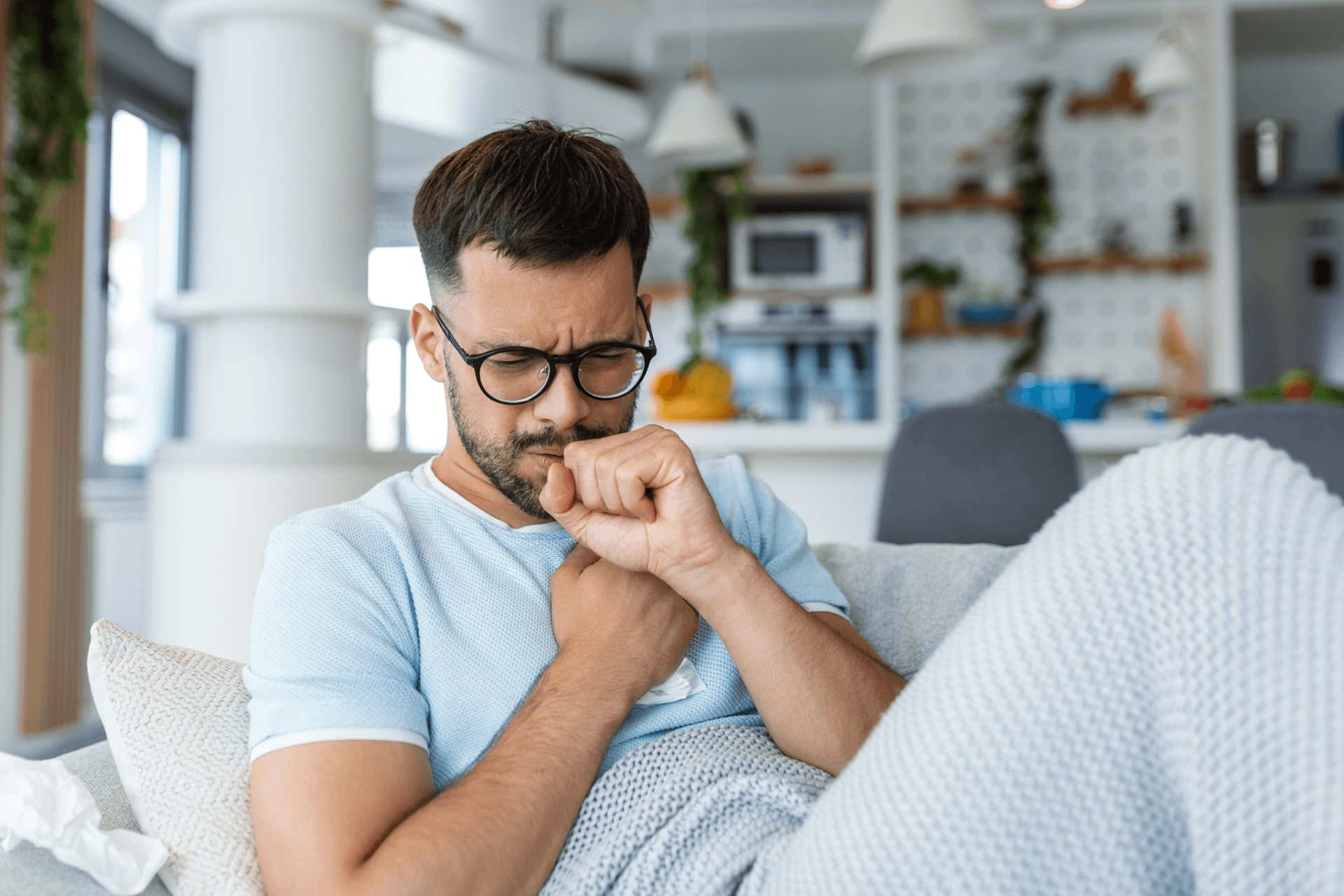 A man in a blue shirt sitting on a couch coughing due to poor winter indoor air quality