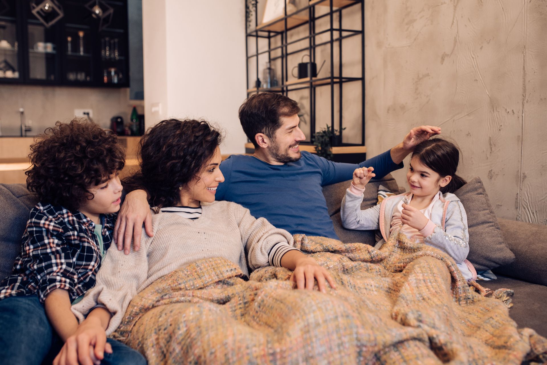 A family of four lounging happily together in their living room, a blanket shared between them